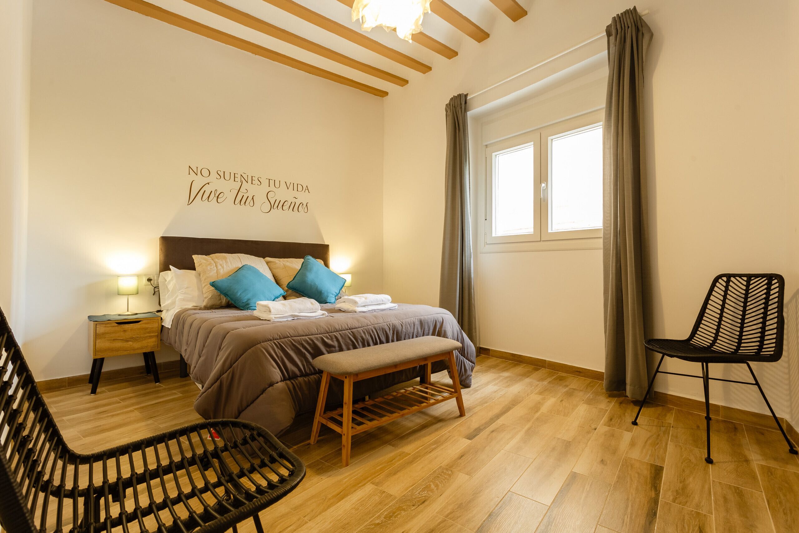 A bedroom with a bed, bedside table, bench, chair, and a window with curtains.