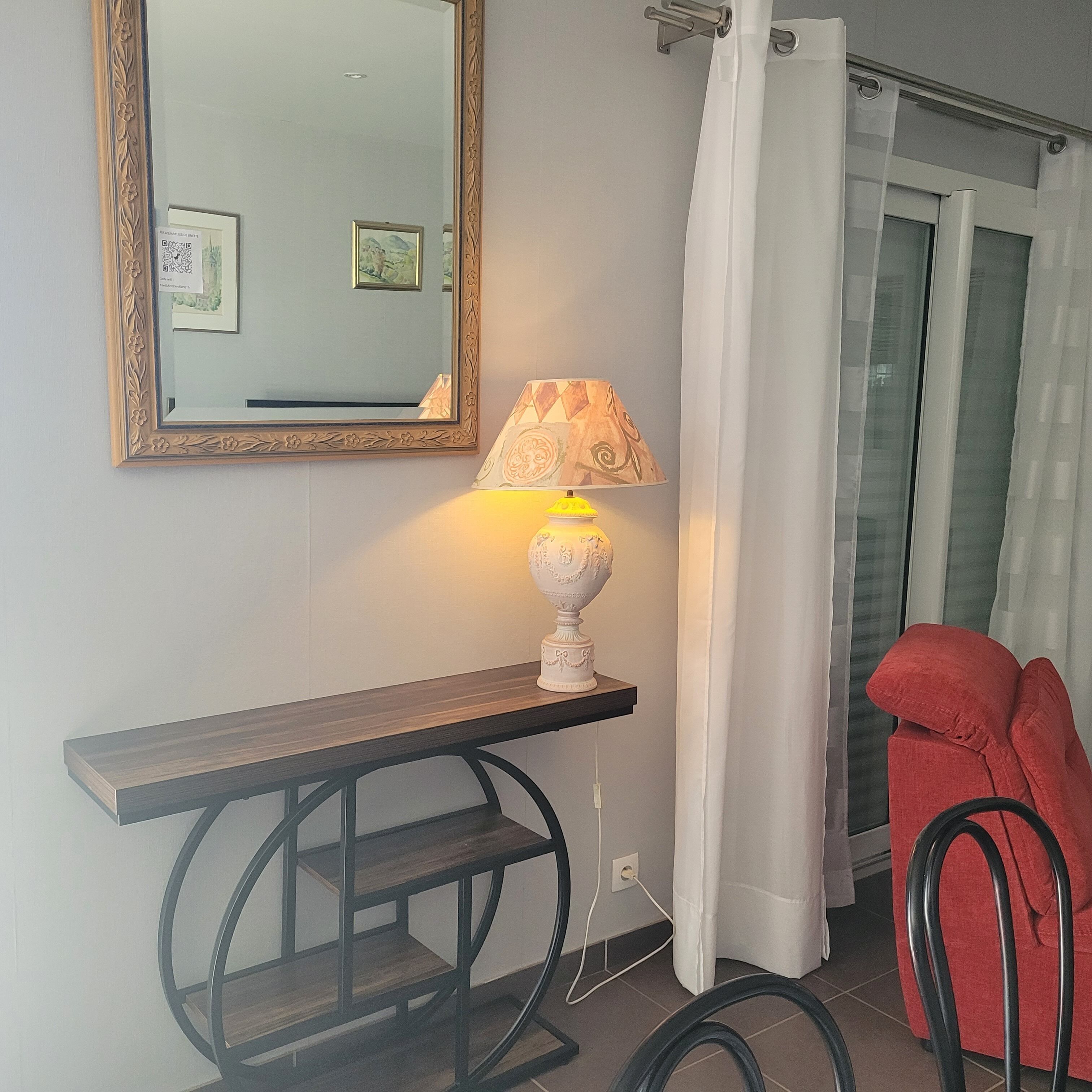 Een kamer met een houten console, een witte lamp, een spiegel met een bloemenversiering en een rode stoel.