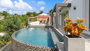 A modern pool area with a curved edge, a covered seating area, and a view of a house in the background. - SUNSET VILLA ANGEL **** (saint barthelemy)