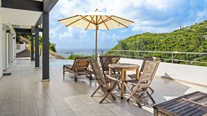 An outdoor patio with wooden furniture and a large umbrella, overlooking a lush green hillside and the ocean. - SUNSET VILLA ANGEL **** (saint barthelemy)