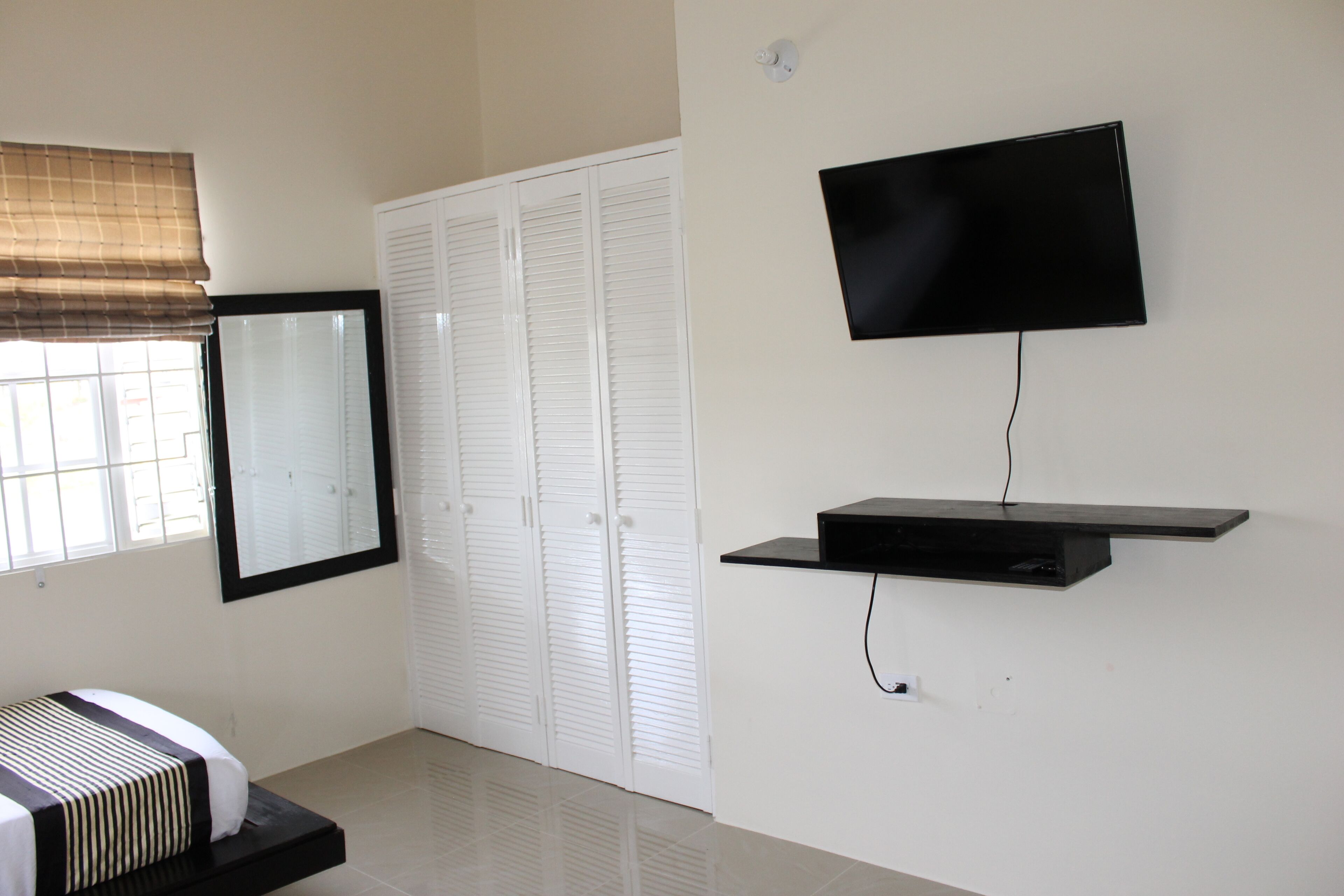 Ein Schlafzimmer mit Bett, Wand-Fernseher, Kleiderschrank und einem Fenster mit Jalousien.