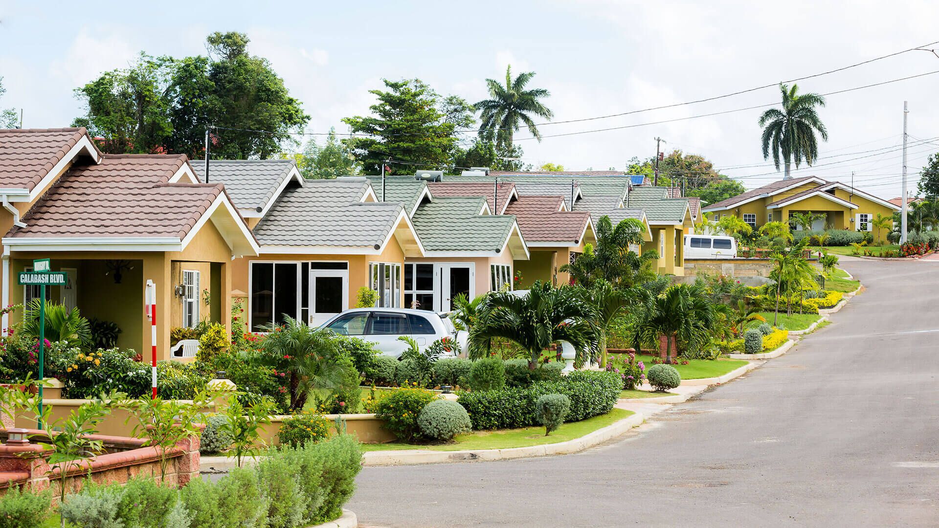 Une rangée de maisons à un étage, dotées de toits inclinés, entourées d’une végétation luxuriante et baignées par un ciel dégagé.