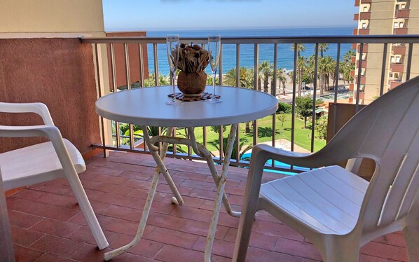 A balcony with a table and chairs, overlooking a view of the sea and buildings.