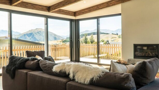 Un salon moderne doté d’un grand canapé d’angle, d’une cheminée et d’une vue imprenable sur les montagnes, grâce à de grandes fenêtres du sol au plafond.