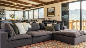 Un salon moderne doté d’un grand canapé d’angle, d’un coin repas avec une table et des chaises, et d’une grande fenêtre offrant une vue sur les montagnes.