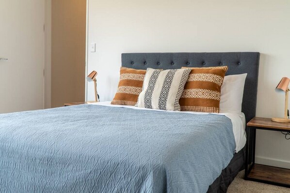 A neatly made bed with a blue textured bedspread, a grey upholstered headboard, and patterned pillows. There are two bedside lamps on wooden nightstands.