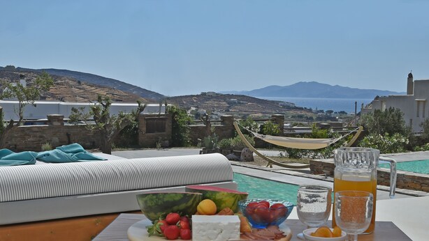 En terrasse med et spisebord dækket med frugt, ost og drikkevarer, med udsigt over en pool og et smukt landskab med bakker og hav.