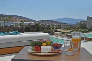 Terraço com mesa posta, com frutas, queijos e bebidas, dando para uma piscina e uma vista panorâmica de colinas e do mar.