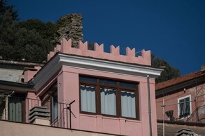 A pink building with a balcony, a window with curtains, and a stone structure in the background.