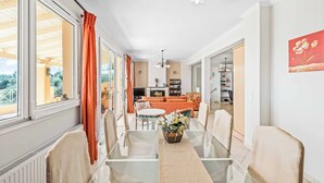 Ein Essbereich mit Glastisch und Stühlen, einer Blumenvase und Blick auf das Wohnzimmer mit Sofa und orangefarbenen Vorhängen.