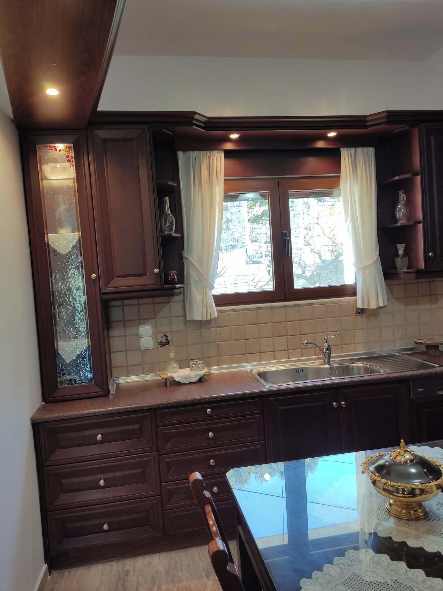A kitchen with dark wooden cabinets, a sink, a window with curtains, and a table with a decorative item.