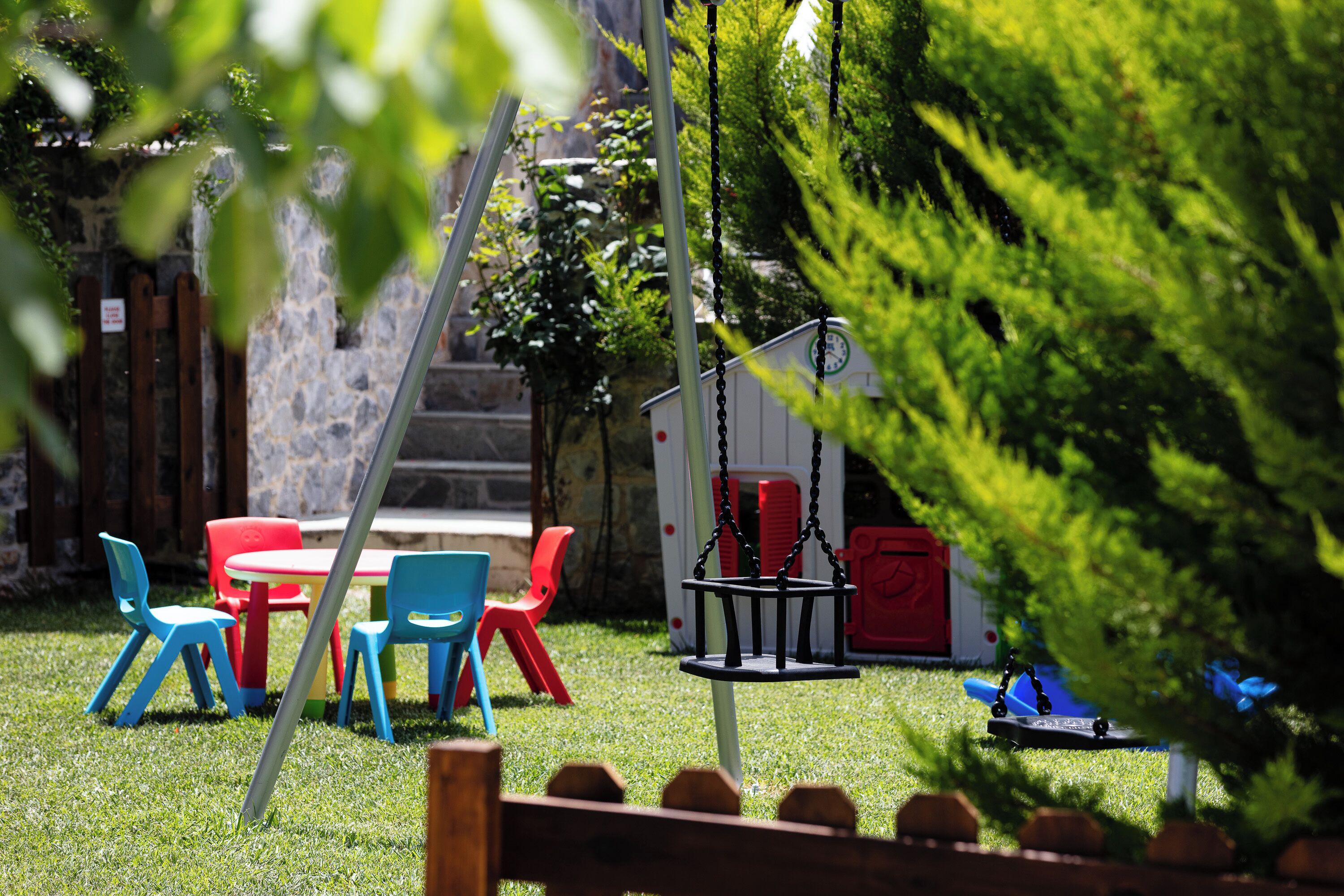 Zona de juegos infantiles con columpios, una mesita y sillas de colores.