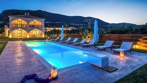 Área da piscina com espreguiçadeiras, um edifício de pedra com arcos e vista para colinas ao fundo.