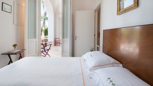 Ein Schlafzimmer mit einem großen Bett, einem hölzernen Kopfteil, einem kleinen Tisch mit einer Vase und einer Tür zu einem Außenbereich mit Stühlen.