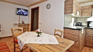 Un comedor con una mesa de madera, sillas y un pequeño jarrón de flores. Hay un televisor montado en la pared y un reloj también en la pared.