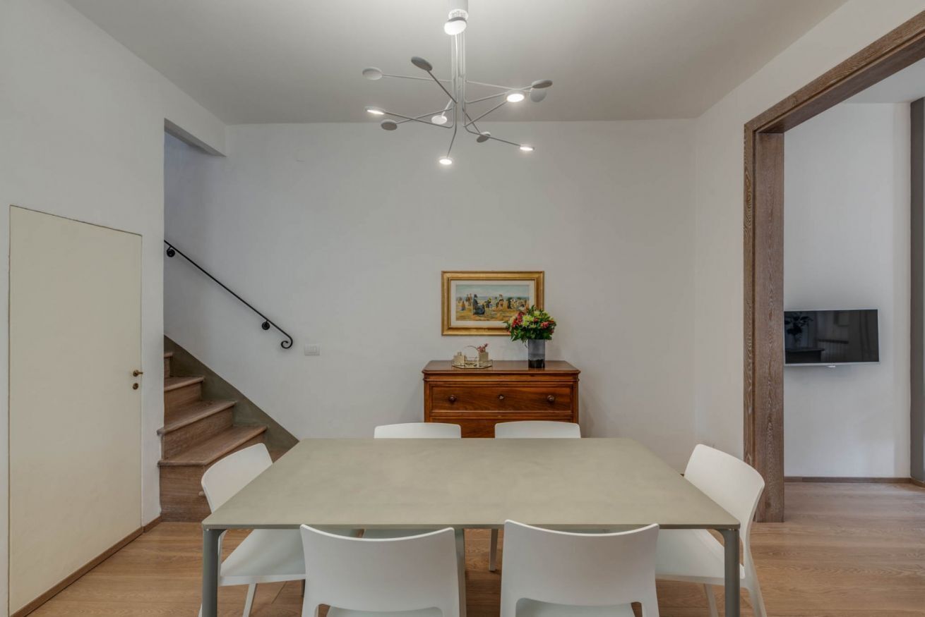 A dining area with a table, chairs, a staircase, a painting, a vase with flowers, and a television on the wall.