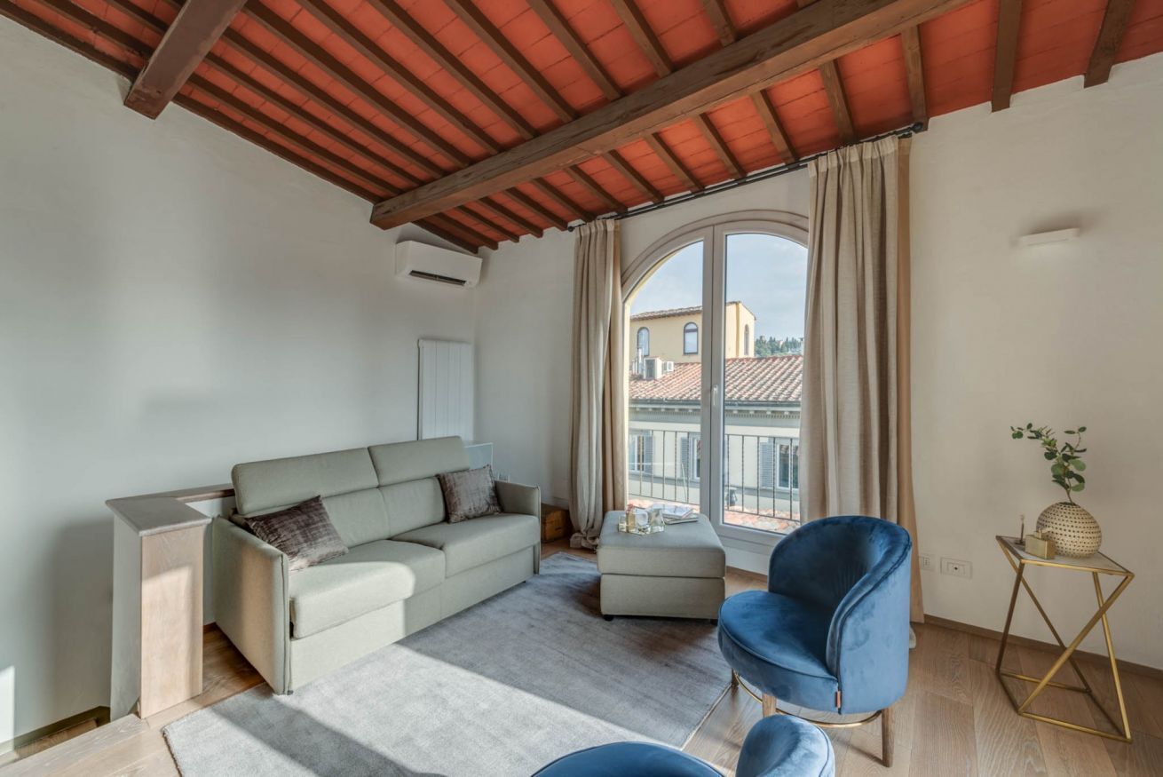 A modern living room with a sofa, ottoman, and blue chair, a wooden side table, and a view through a large window.
