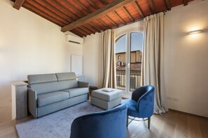 Een moderne woonkamer met een bank, poef en stoel, een groot raam met gordijnen, en een houten plafond.