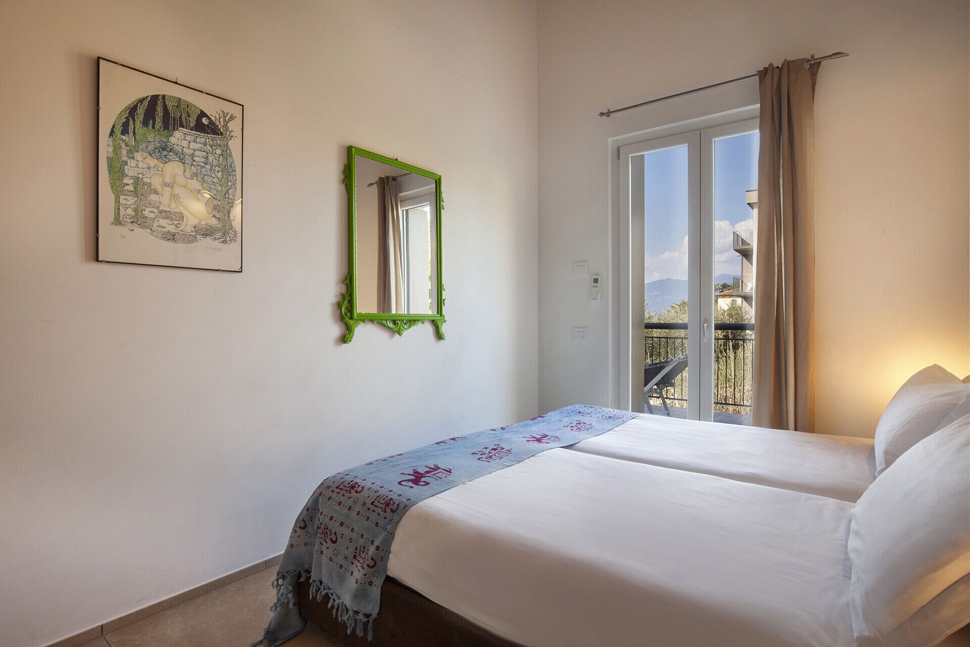 A bedroom with a bed, a green-framed mirror, a framed picture, and a balcony with a view.