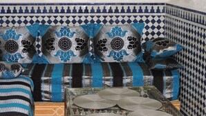 Un sofá de estilo marroquí tradicional con intrincados diseños azules y plateados, una mesa de centro de cristal con platos decorativos y una pared de azulejos con motivos geométricos.