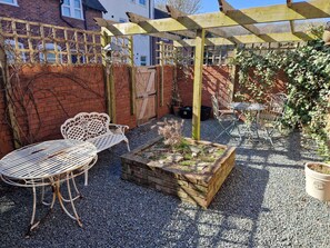 Un espace patio agrémenté d’une pergola, d’une table et d’une chaise en métal blanc, d’un bac à fleurs en bois et d’un mur en briques.
