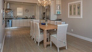 A dining area with a wooden table and wicker chairs, a chandelier, and a kitchen in the background.