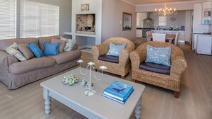 A living room with a beige sofa, two wicker chairs, a coffee table with a book and candle, and a kitchen in the background.