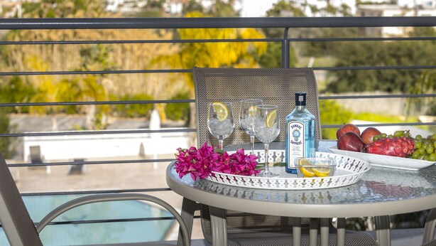 A round table with a bottle, glasses, fruit, and flowers on a balcony overlooking a pool and buildings. - Protaras Palm Suite 207 (Protaras)