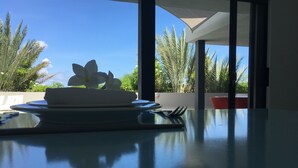 Una mesa de comedor con un plato de flores y un teléfono inteligente, una silla roja, y una vista de palmeras y cielo despejado a través de un ventanal.