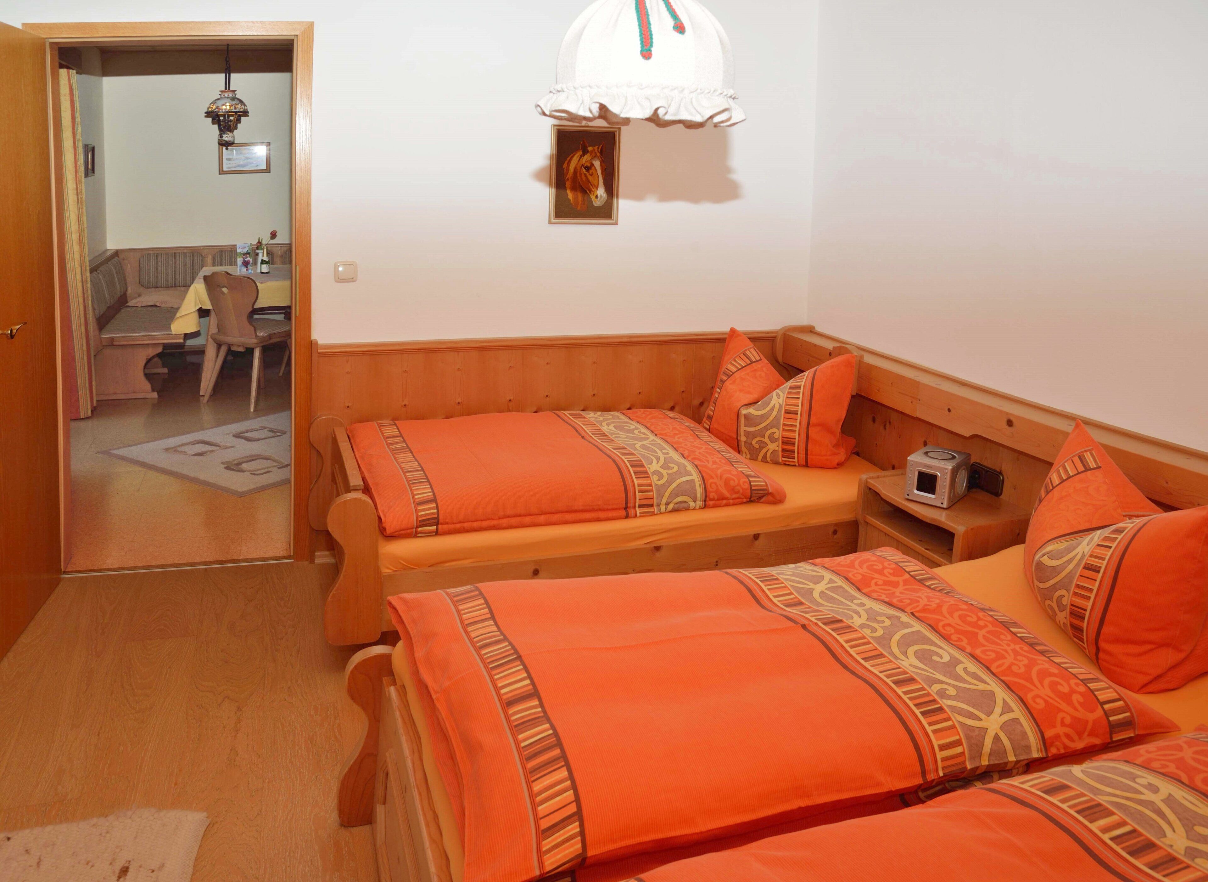 Een kamer met twee houten bedden met oranje beddengoed, een nachtkastje van hout met een camera, en een witte lamp aan de muur.