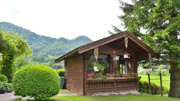 A wooden cabin with a porch, surrounded by lush greenery and mountains in the background. - No.4 Apartment 50 sqm with one separate bedroom, kitchen-living room, east-facing balcony (Aschau im Chiemgau)