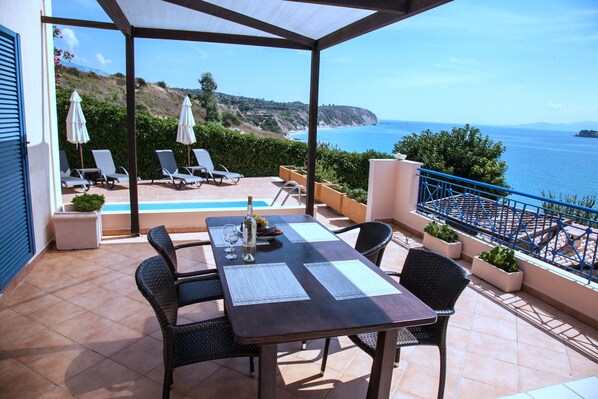 A covered outdoor seating area with a table, chairs, and umbrellas, overlooking a coastal landscape.