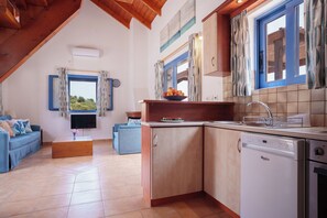 A kitchen with a dishwasher, a sink, and a window with curtains. There is a blue sofa and a wooden coffee table in the living area.