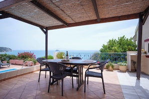 Un patio exterior techado con mesa y sillas, una piscina y vista al mar.