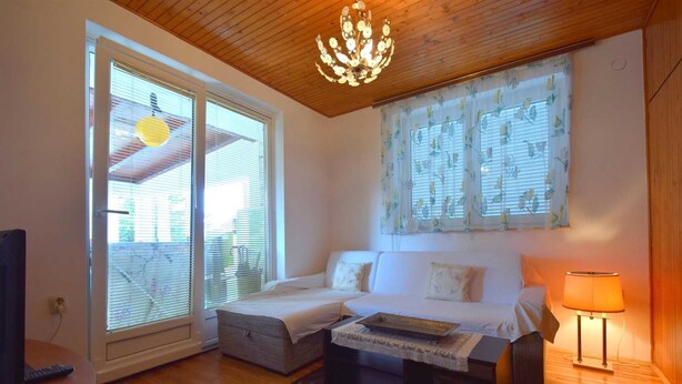 Una sala de estar acogedora con un sofá, una mesita y una lámpara. Cuenta con ventanas grandes con persianas y una lámpara de techo.