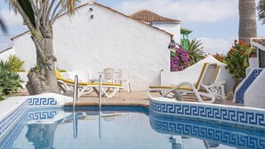 Una piscina con mosaicos azules, rodeada de sillones blancos y una pared blanca con techo de tejas.