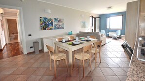 Amplia sala de estar con una mesa de comedor, sillas y un sofá. Se observan pinturas abstractas en las paredes, una ventana que proporciona luz natural y una cocina visible al fondo.