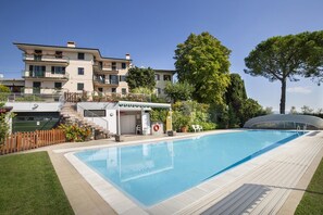 A swimming pool with a wooden deck, a covered seating area, and a building with balconies in the background. - Apartment Montegolo Four With Pool And Lake View (Costermano)