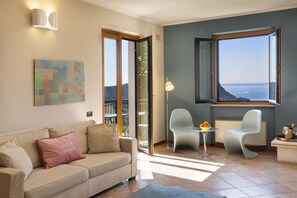 A living room with a sofa, chairs, a table with fruit, and a view of the sea.