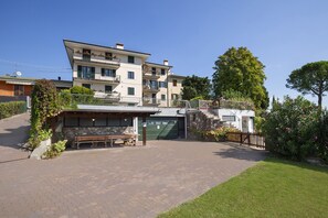 A multi-story residential building with a paved driveway, a covered outdoor seating area, and a stone wall. - Apartment Montegolo Four With Pool And Lake View (Costermano)