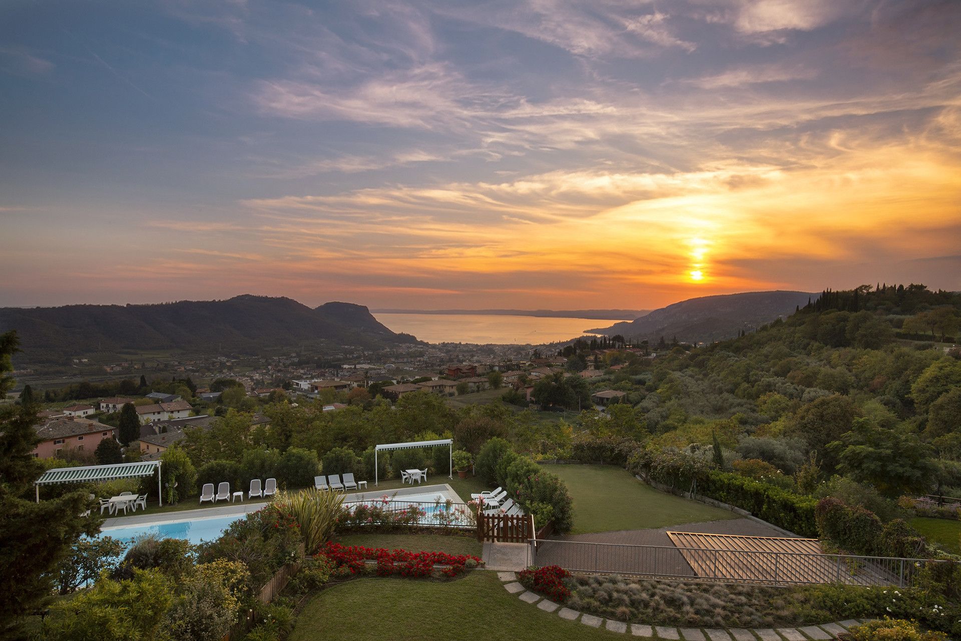 A residential area with a swimming pool, garden, and a view of the sunset over a body of water and hills.