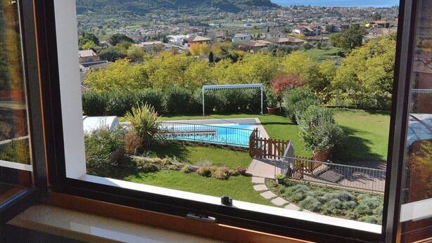 Vista desde una ventana que muestra una zona residencial con piscina, césped verde y colinas al fondo.