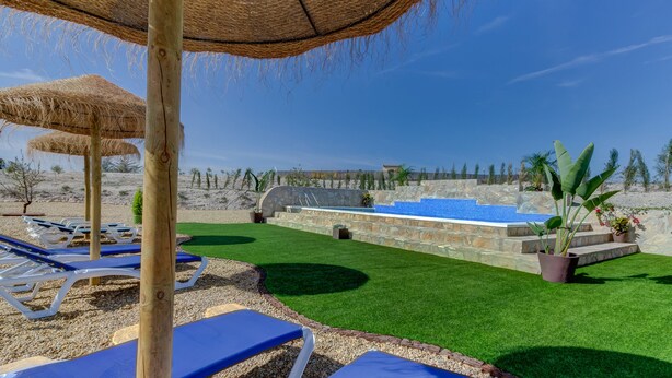 A pool area with blue lounge chairs and thatched umbrellas, surrounded by a well-manicured lawn and stone walls. - Villa Damara, luxury apartment Oliva, the place to relax! (Albox)