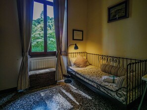 Une petite pièce agréable, avec un lit simple, une fenêtre avec des rideaux, un radiateur et un tableau encadré au mur.