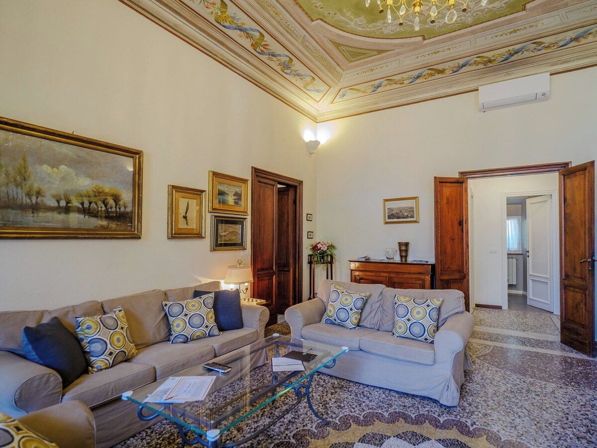 A living room with two sofas, a coffee table, and a chandelier. There are framed pictures on the walls and a door leading to another room.