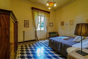 Una habitación de hotel con piso a cuadros, una cama con colcha azul, una silla, una mesa y una ventana con cortinas.