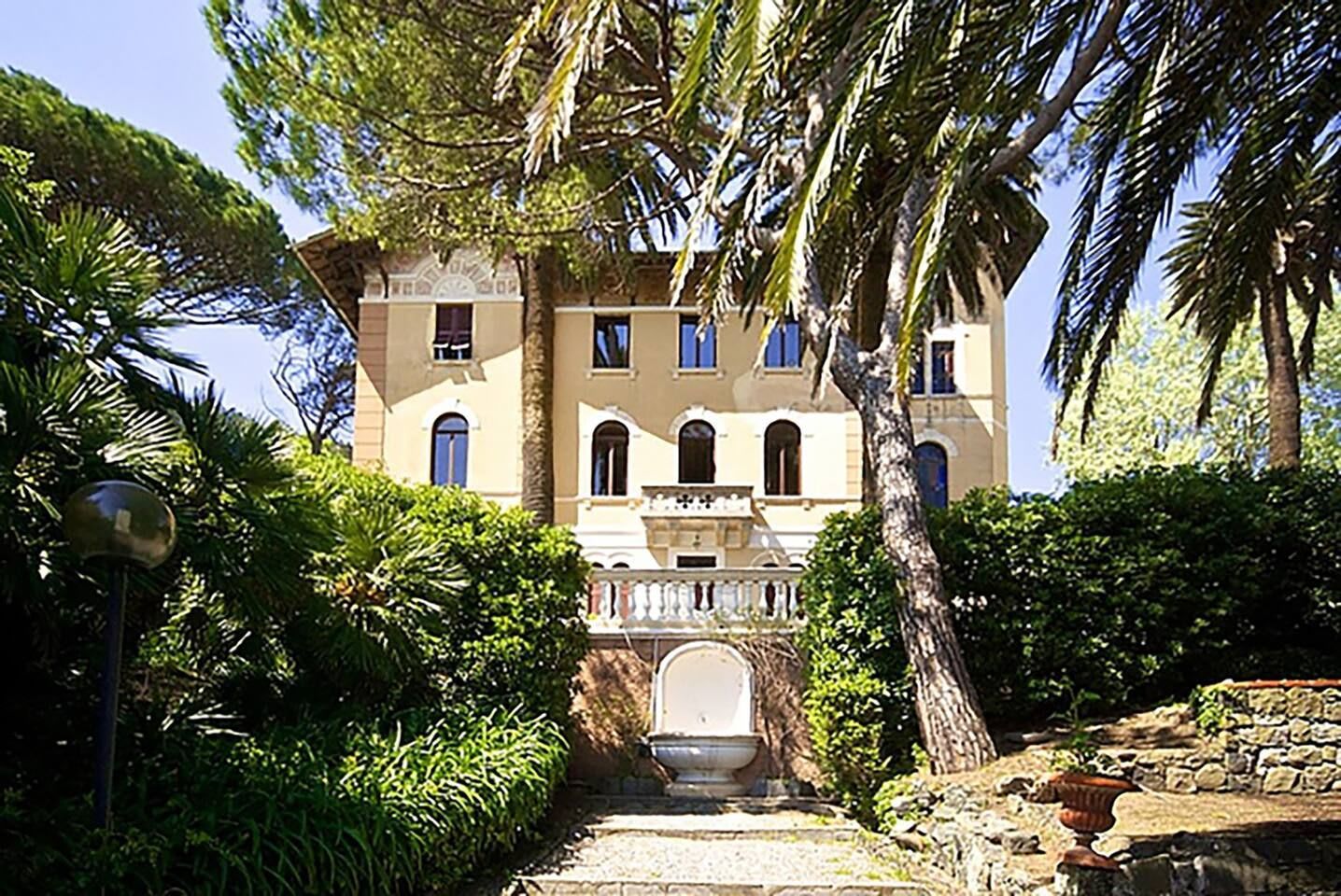 Un grand immeuble à plusieurs étages, doté d’une façade symétrique, d’une entrée centrale surmontée d’un arc décoratif et entouré d’un jardin planté de palmiers et d’arbustes.