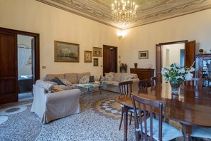 Una sala de estar con un sofá, una mesa de comedor y sillas. Hay cuadros en la pared y una araña.