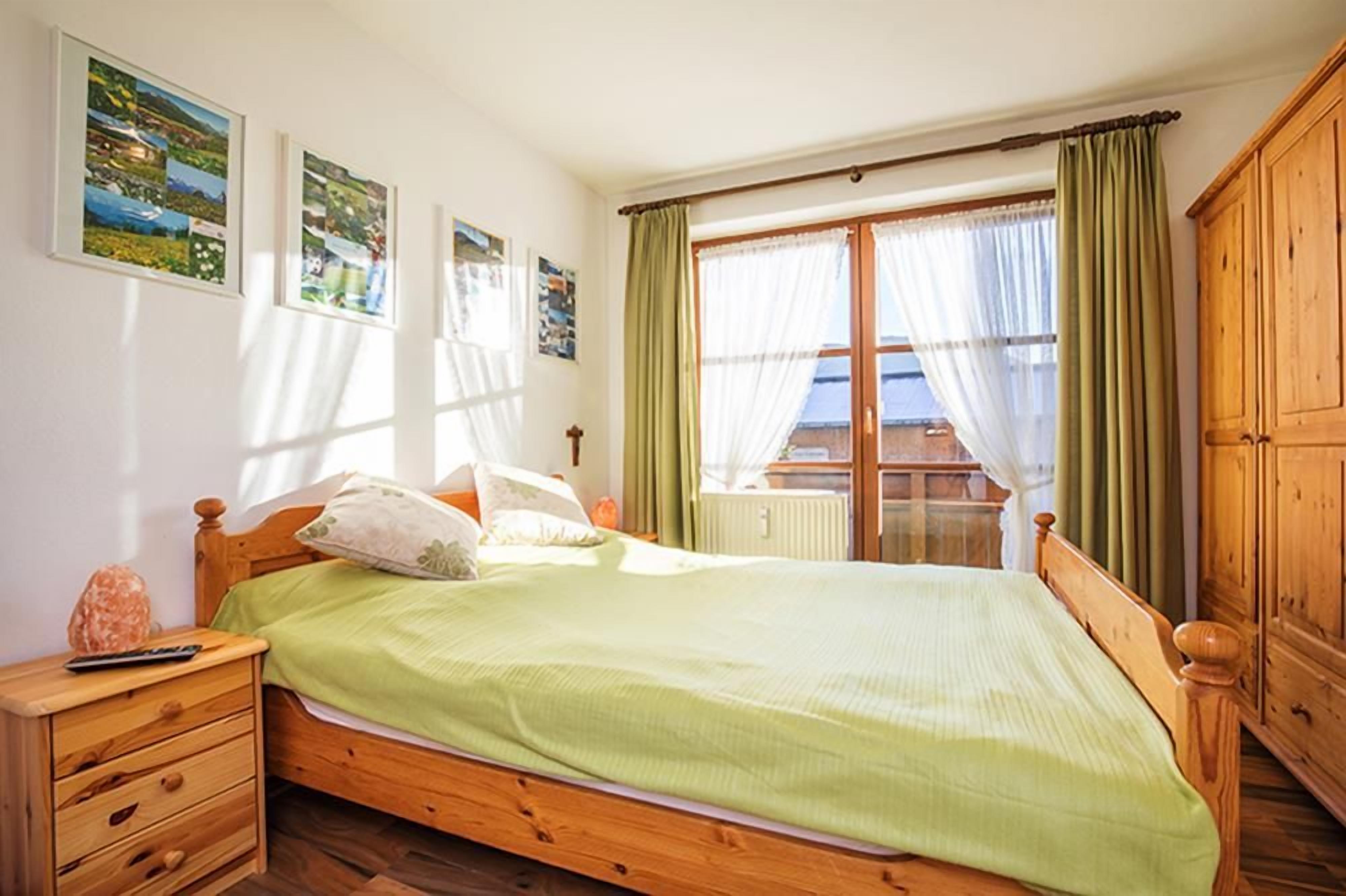 A bedroom with a wooden bed, a nightstand, a window with curtains, and a view of the outside.