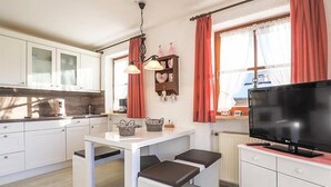 Uma cozinha compacta com armários brancos, uma mesa de jantar com bancos, uma televisão e uma janela com cortinas vermelhas.